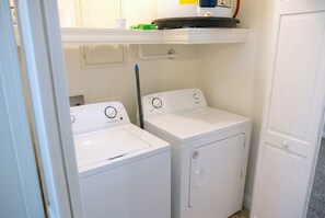 Un lavadero con dos lavadoras blancas, una secadora y un estante superior con un contenedor verde y una taza blanca.
