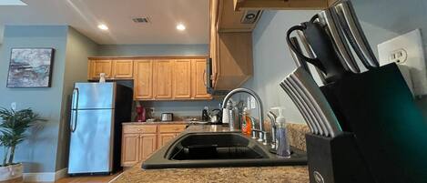 A kitchen with wooden cabinets, a stainless steel refrigerator, a granite countertop, a sink with a faucet, and a black appliance on the right.