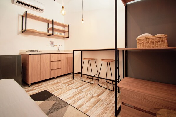 A compact kitchen with wooden cabinets, a countertop, and a sink. There are two stools under a shelf, and a basket on another shelf. - Lexum by Bernalo Hotels (Medellín)