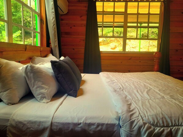 A wooden cabin room with a bed, pillows, and a window with a view of greenery.
