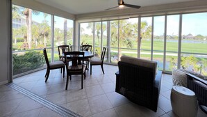 Una veranda soleggiata con tavolo e sedie, una ventola a soffitto e ampie finestre che si affacciano su un campo da golf e palme.