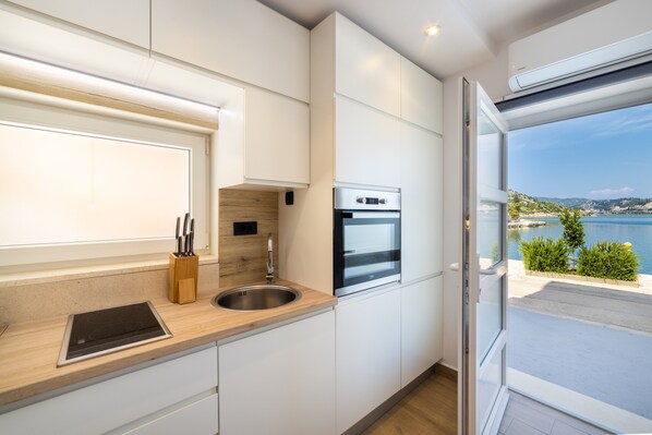 A modern kitchen with a built-in oven, a sink, and a window overlooking a scenic coastal view. - Apartments Ema - Studio Apartment with Sea View (Rogotin)