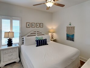 A bedroom with a bed, a ceiling fan, a bedside table with a lamp, and a wall-mounted macrame decoration. - Sun Kissed Memories by Southern Vacation Rentals (Panama City Beach)