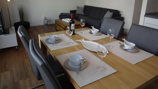 A dining area with a wooden table set for four, a sofa, and a wall-mounted compass. - Ter/101 - Terrassenhuegel -teilseeblick (Scharbeutz)