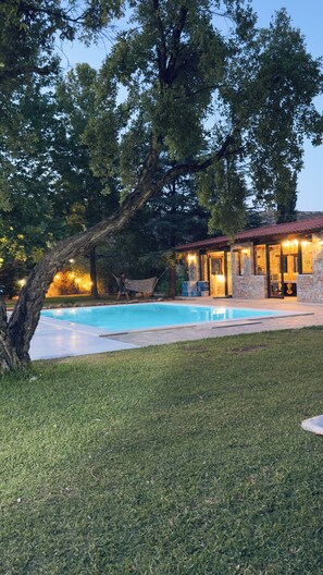 A swimming pool surrounded by a grassy area with a stone building featuring a red-tiled roof and glass doors.