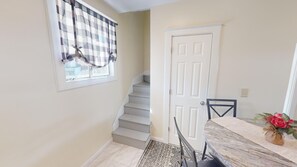 Un escalier menant à une porte, une table à manger avec des chaises et une fenêtre ornée d’un rideau à carreaux.