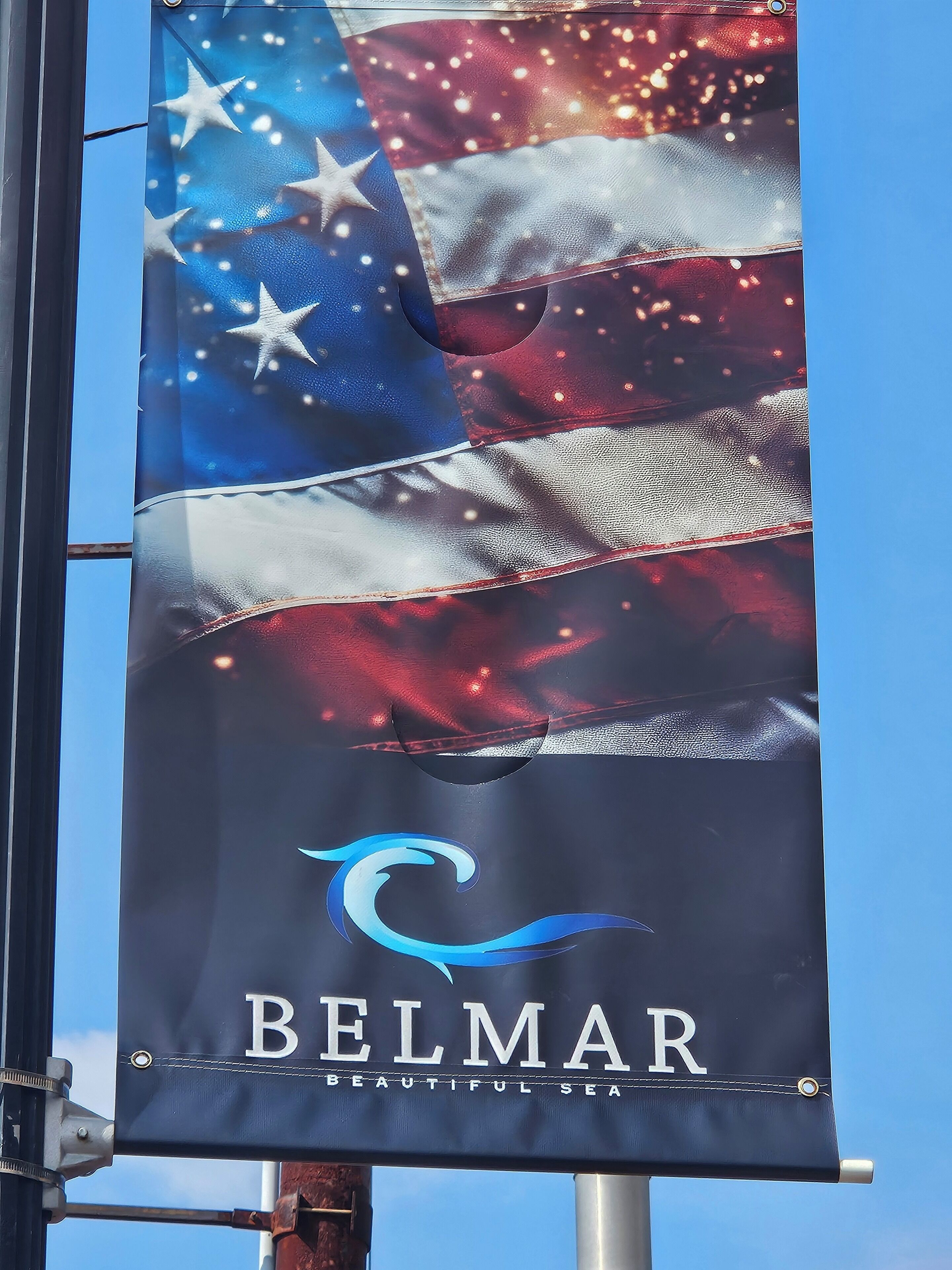 A flag with the Belmar logo and the text 'BELMAR BEAUTIFUL SEA' against a clear sky.