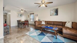 A living room with a brown leather sofa, a dining table with chairs, a ceiling fan, and a blue and white patterned rug.