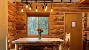 A rustic wooden dining area with a large table and benches, a window with a view of trees, and a ceiling light fixture.