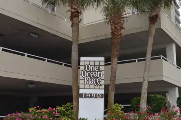 A building with a sign that reads "One Ocean Place 1990" surrounded by palm trees and flowering plants.