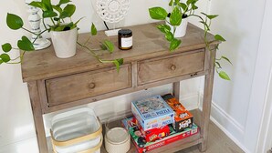 Une console en bois ornée de deux plantes en pot, d’une bougie et d’une assiette décorative. Sous la console, on trouve des paniers de rangement et une boîte de jeu de Monopoly.