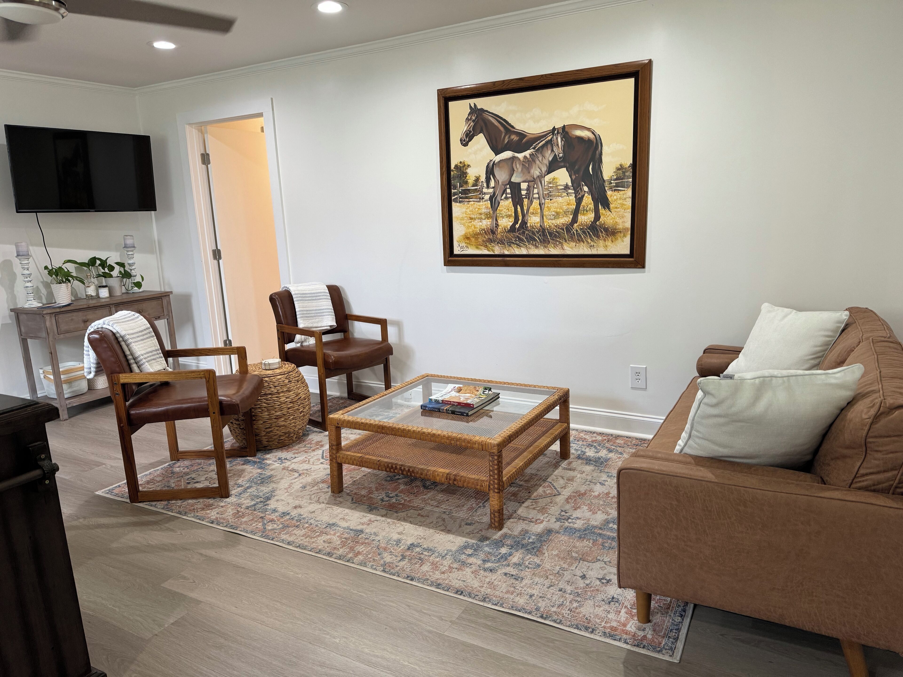 Sala de estar com um sofá de couro marrom, uma mesa de centro de vime e duas poltronas de couro. Um quadro com cavalos está pendurado na parede.