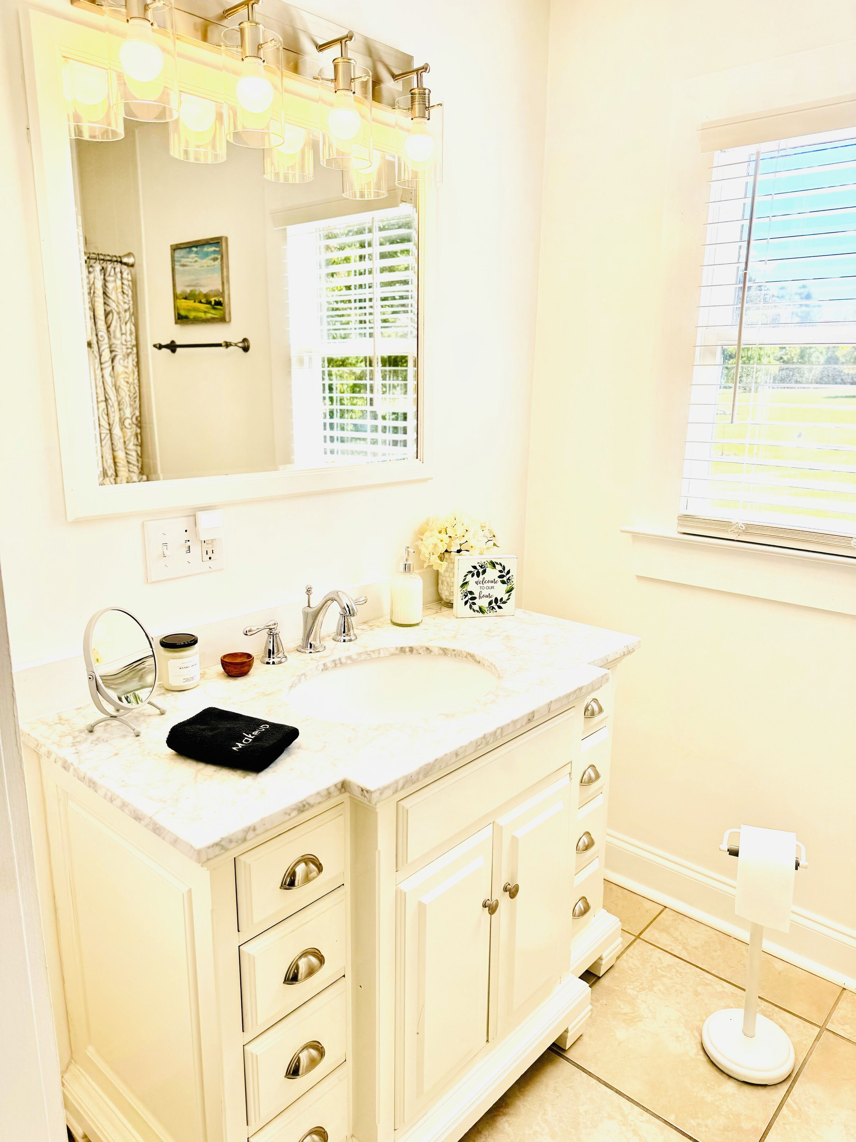 A bathroom with a white vanity, a large mirror, a sink, and a window with blinds.