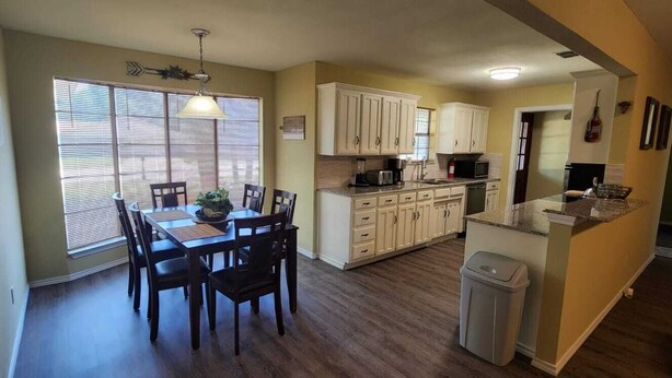 A kitchen with wooden cabinets, a dining table with chairs, a trash can, and a window with blinds.