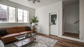 A living room with a brown leather sofa, a glass coffee table, a patterned rug, and a door leading to a staircase.