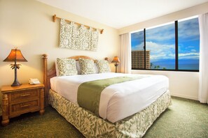 Habitación de hotel con una cama grande, dos lámparas de noche, un cabecero estampado, vista al océano y una ventana con cortinas.