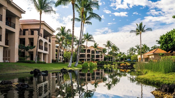 A hotel with multiple floors, a courtyard with palm trees, and a pond reflecting the surroundings.