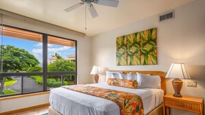 A hotel room with a large window, a bed with floral bedding, two bedside lamps, and a ceiling fan.