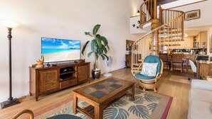 Un salón amplio con una mesa de centro de madera, un televisor, una planta grande en maceta y una silla de mimbre.
