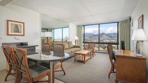 A hotel room with a dining area, a sofa, a desk, and a large window offering a city and mountain view. - Aston Waikiki Sunset 1BMV | Mountain Views – Heated Pool & Spa (Honolulu)