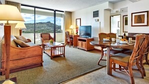 A hotel room with a large window, a television, a dining area, and a seating area with a view of the mountains. - Aston Waikiki Sunset 1BMV | Mountain Views – Heated Pool & Spa (Honolulu)