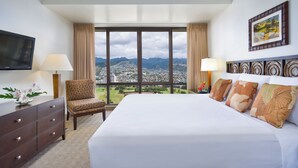 A hotel room with a large bed, a chair, a TV, and a view of a city and mountains. - Aston Waikiki Sunset 1BMV | Mountain Views – Heated Pool & Spa (Honolulu)