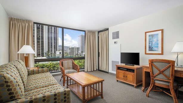 Habitación de hotel con sofá, dos sillones, televisor, escritorio y ventana con vistas a la ciudad.