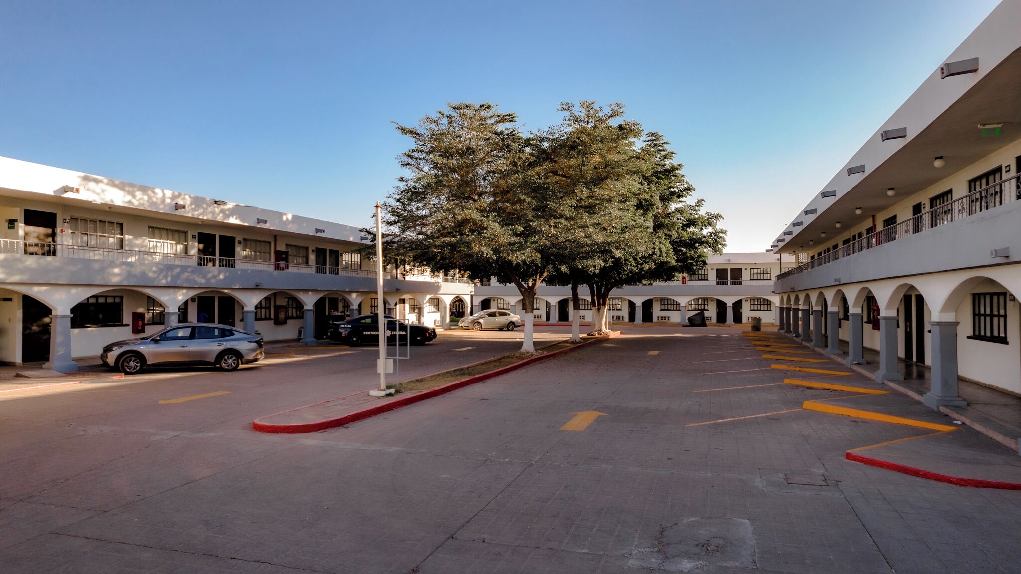 Un patio de hotel con un árbol central, varios espacios de estacionamiento y un edificio con ventanas y balcones arqueados.