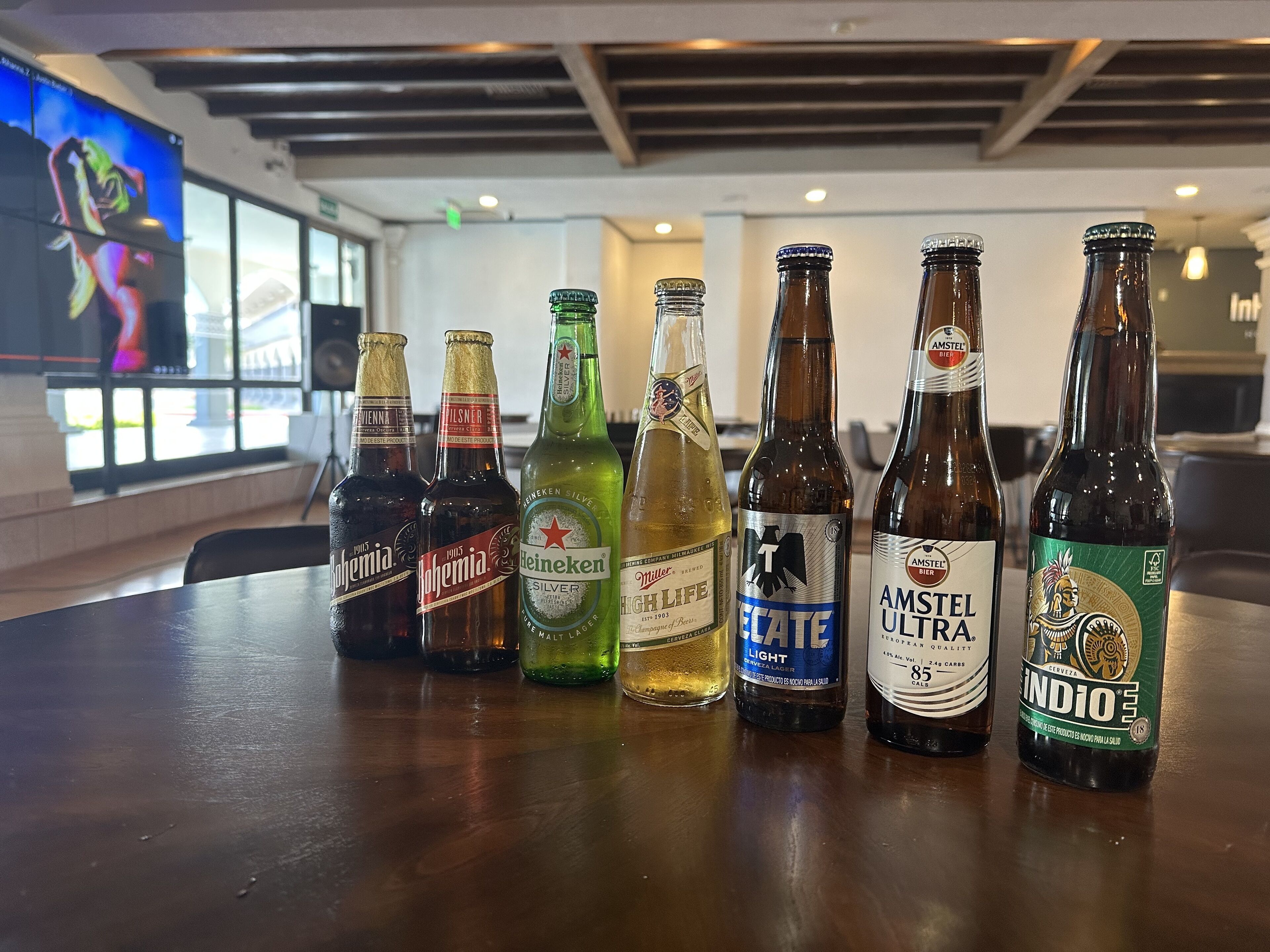 A row of beer bottles on a table in a modern interior with large windows and a visible TV screen.