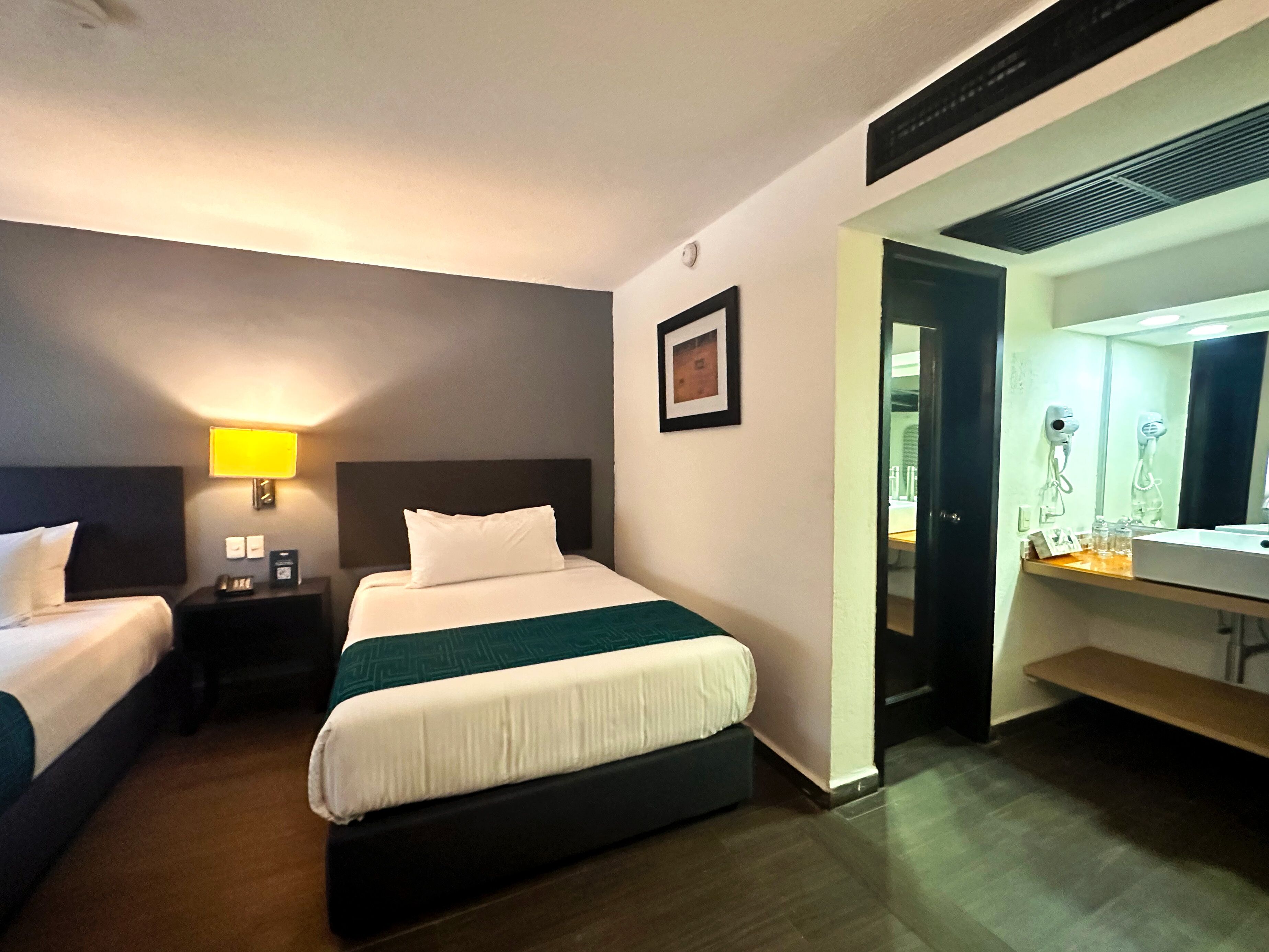 Habitación de hotel con dos camas, una mesita de noche con lámpara, un cuadro enmarcado en la pared y un baño con espejo y secador de pelo.