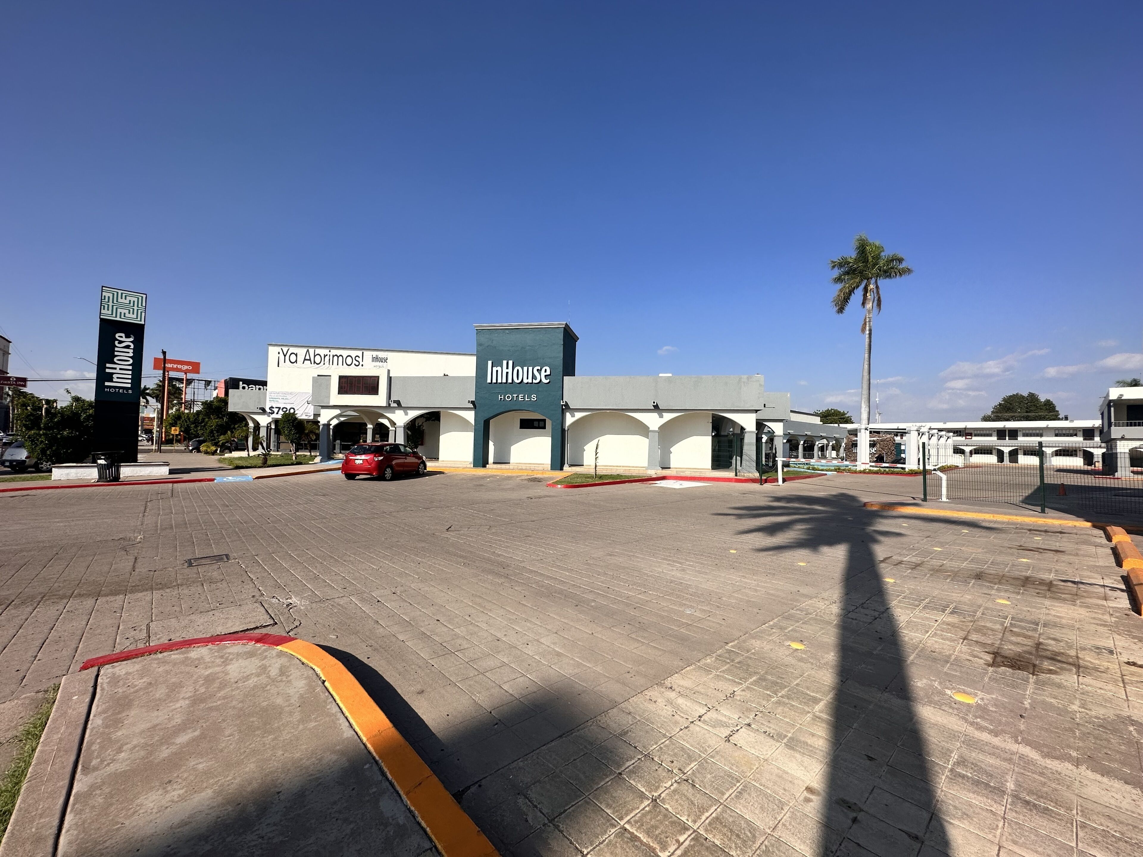 A hotel entrance with a clear sky, a red car parked in front, and a palm tree to the right.