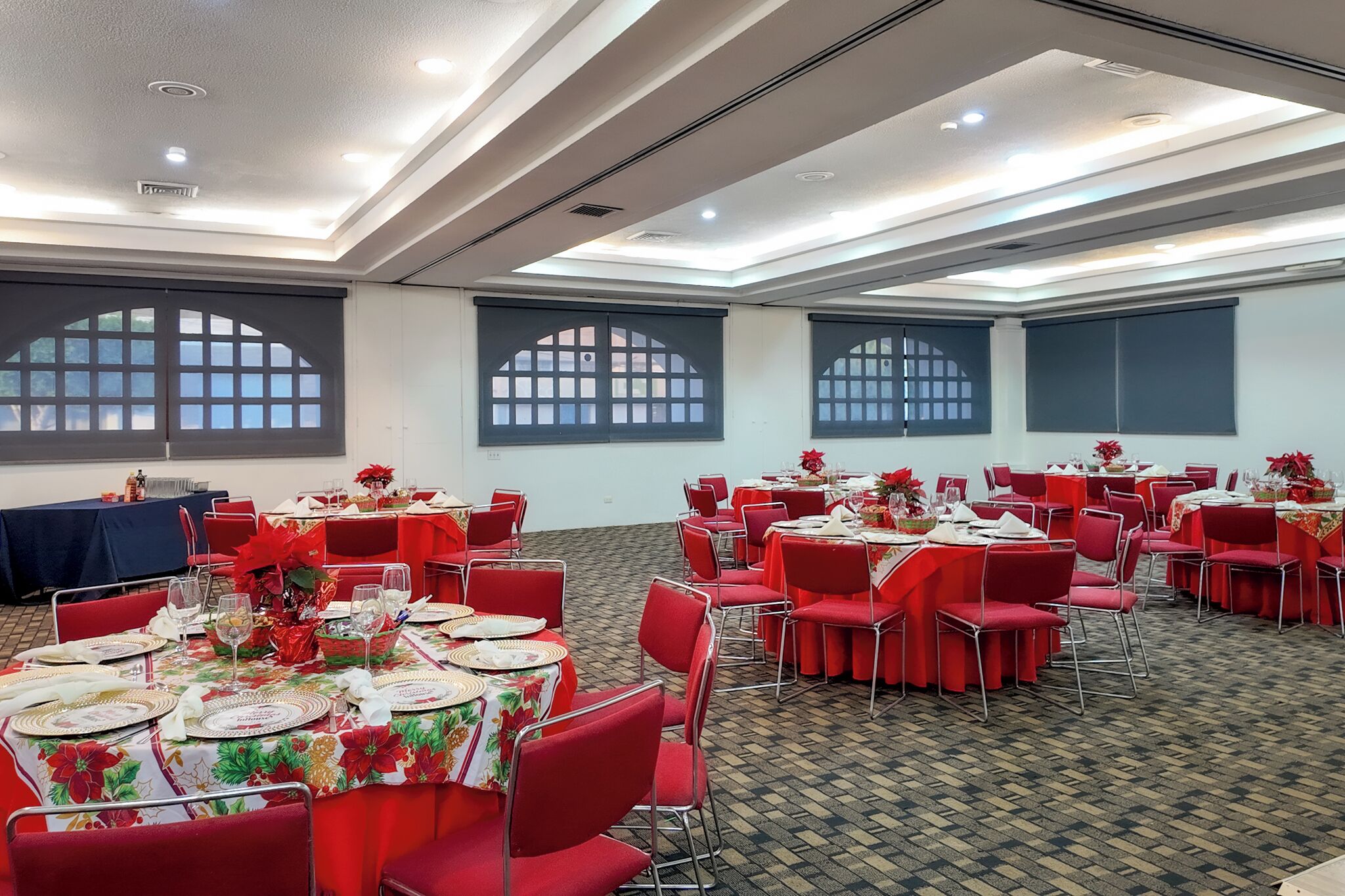 Un salón de banquetes con mesas redondas dispuestas para el servicio de comidas, sillas rojas y amplias ventanas con arcos.