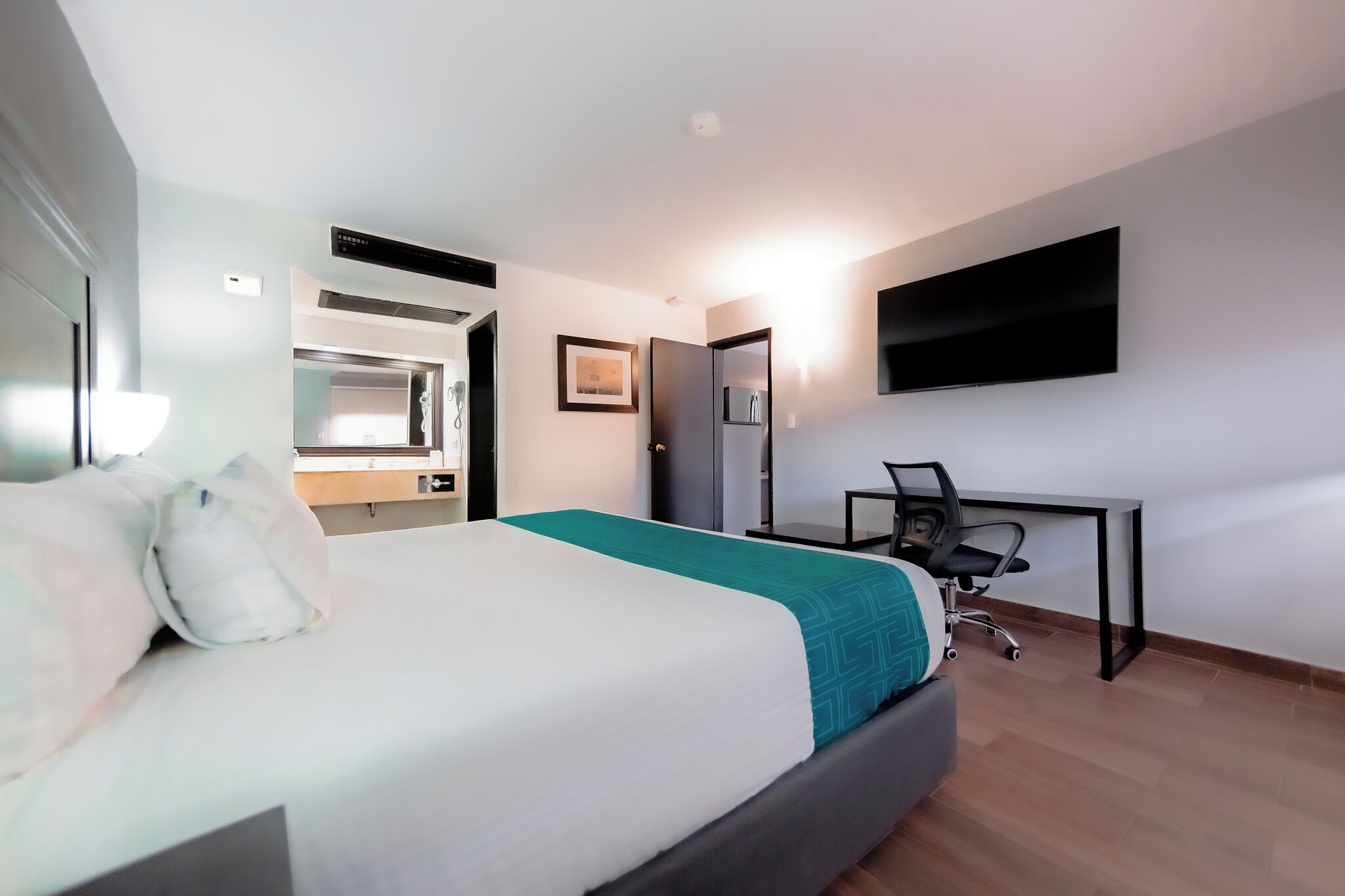 A hotel room with a bed, desk, chair, TV, and a bathroom with a mirror and sink.