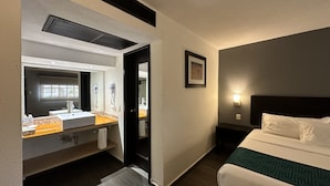 Une chambre d’hôtel comprenant un lit, une table de chevet, une commode, un miroir, un lavabo et un porte-serviettes.
