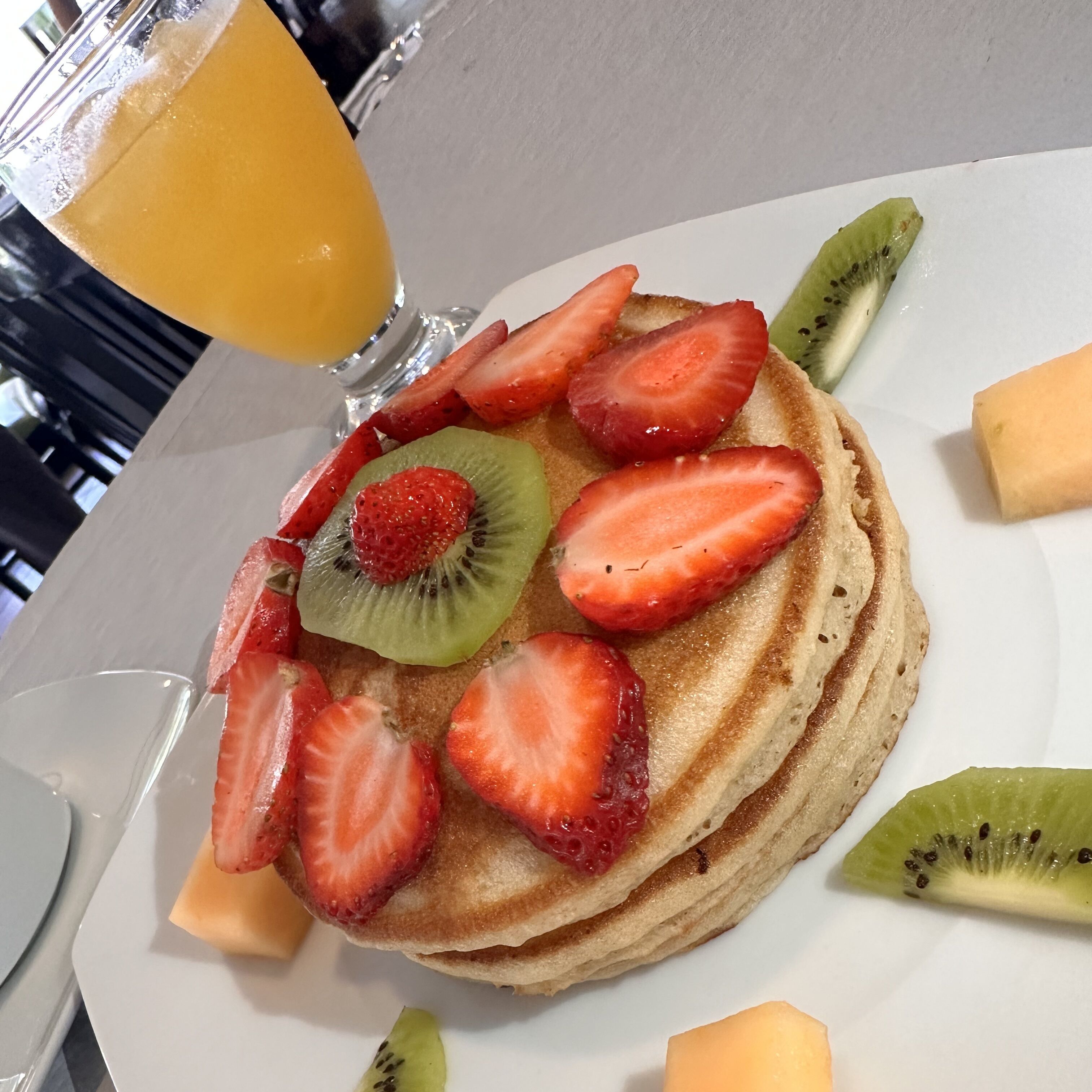 Una pila de panqueques cubiertos de fresas y rodajas de kiwi, acompañada de un vaso de jugo de naranja.