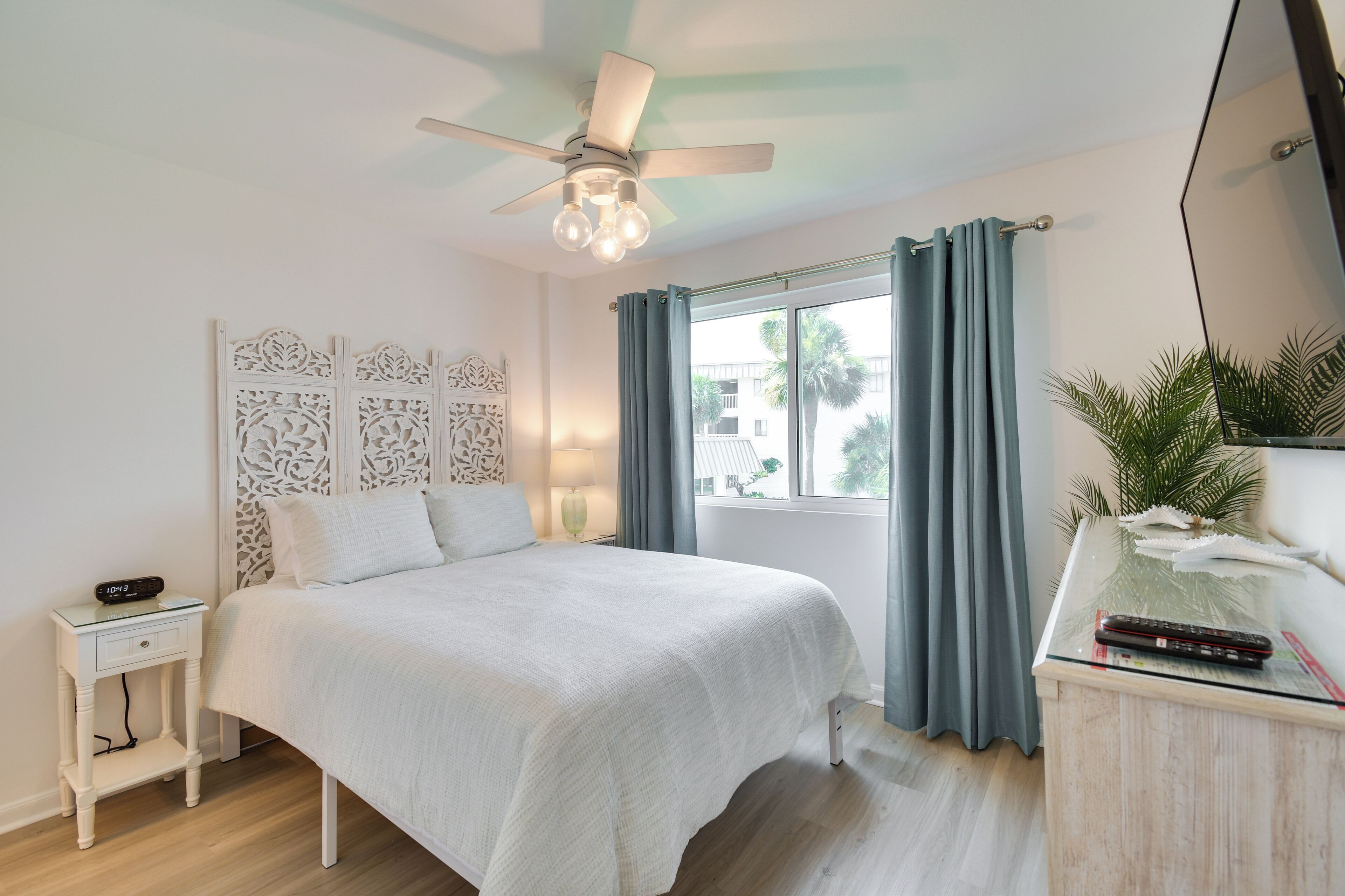 Un dormitorio con cama, mesita de noche, escritorio, ventilador de techo y ventana con cortinas.