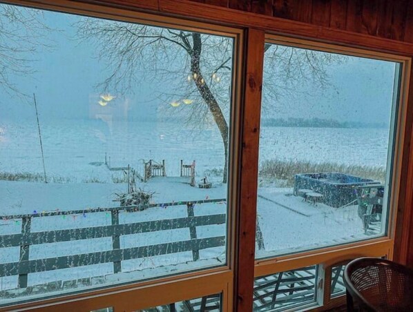 A view through a window of a wooden cabin with a snow-covered deck and a lake.