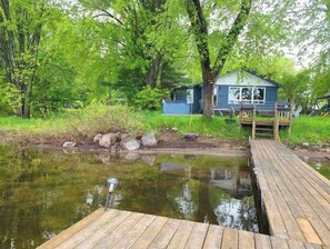 A wooden dock extending over a calm body of water, surrounded by lush greenery and a small cabin with a deck.