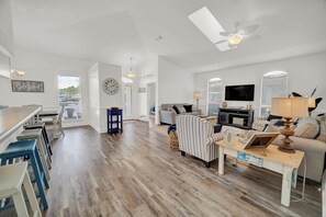 A spacious living room with a large TV, a dining area with bar stools, and a kitchen in the background.