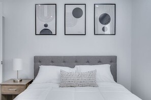 Um quarto com cabeceira estofada cinza, roupa de cama branca, abajur e três obras de arte abstratas emolduradas na parede.