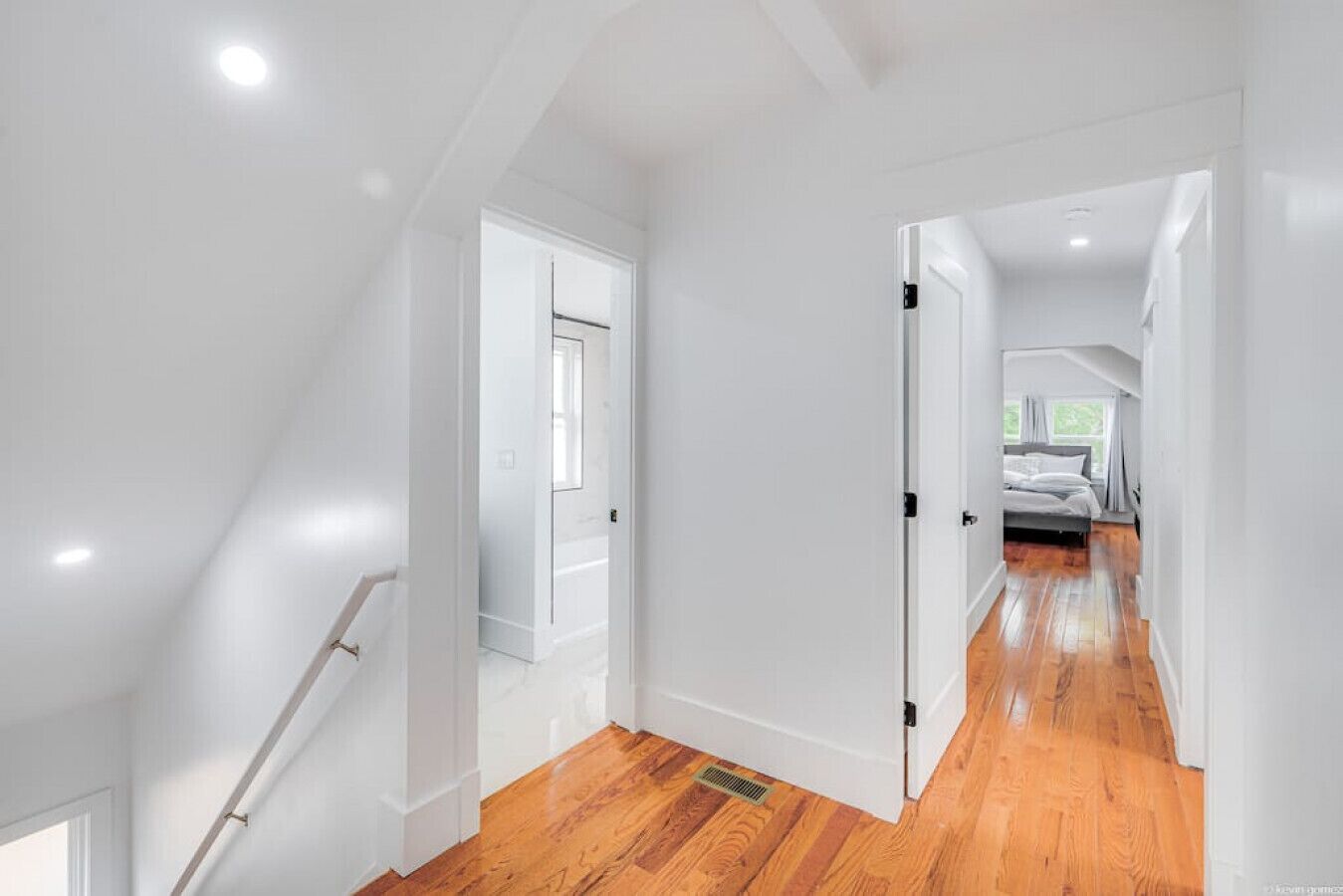A modern, well-lit hallway with wooden flooring and white walls, leading to a bedroom with a bed and a window.