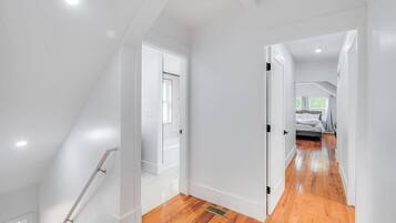 A modern, well-lit hallway with wooden flooring and white walls, leading to a bedroom with a bed and a window.