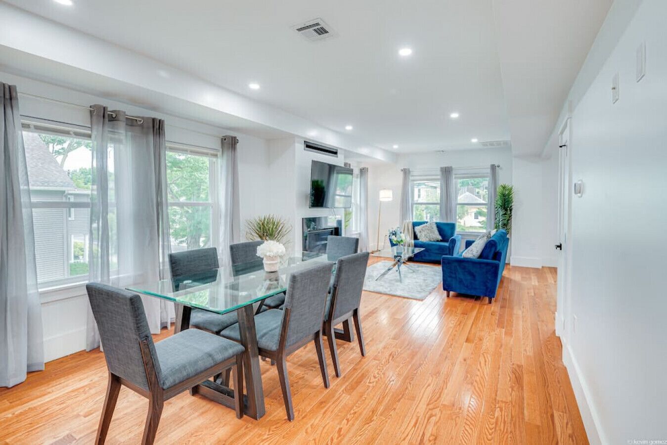 A modern living room with a glass dining table, grey chairs, a blue sofa, and a TV mounted on the wall.
