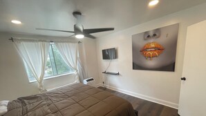 Un dormitorio con cama, ventilador de techo, televisor montado en la pared y ventana con cortinas.