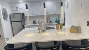 Una cocina moderna con encimera de mármol, tres taburetes oscuros de azul, armarios blancos y luces colgantes.