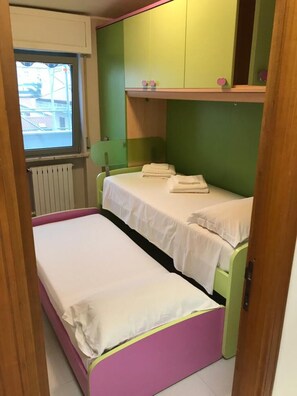 Une petite chambre avec un lit simple, aux murs peints en vert et en rose, une fenêtre donnant sur des immeubles et un radiateur.