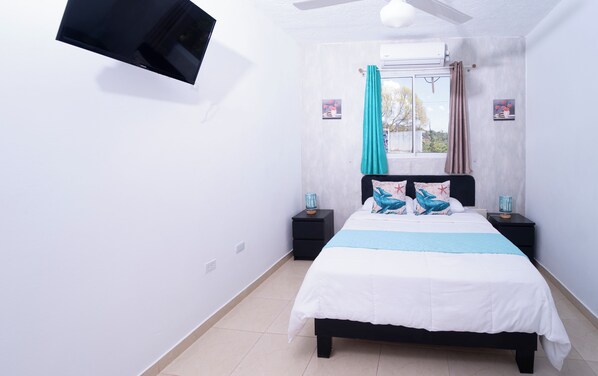 A bedroom with a bed, nightstands, a TV mounted on the wall, an air conditioner, and a window with curtains.