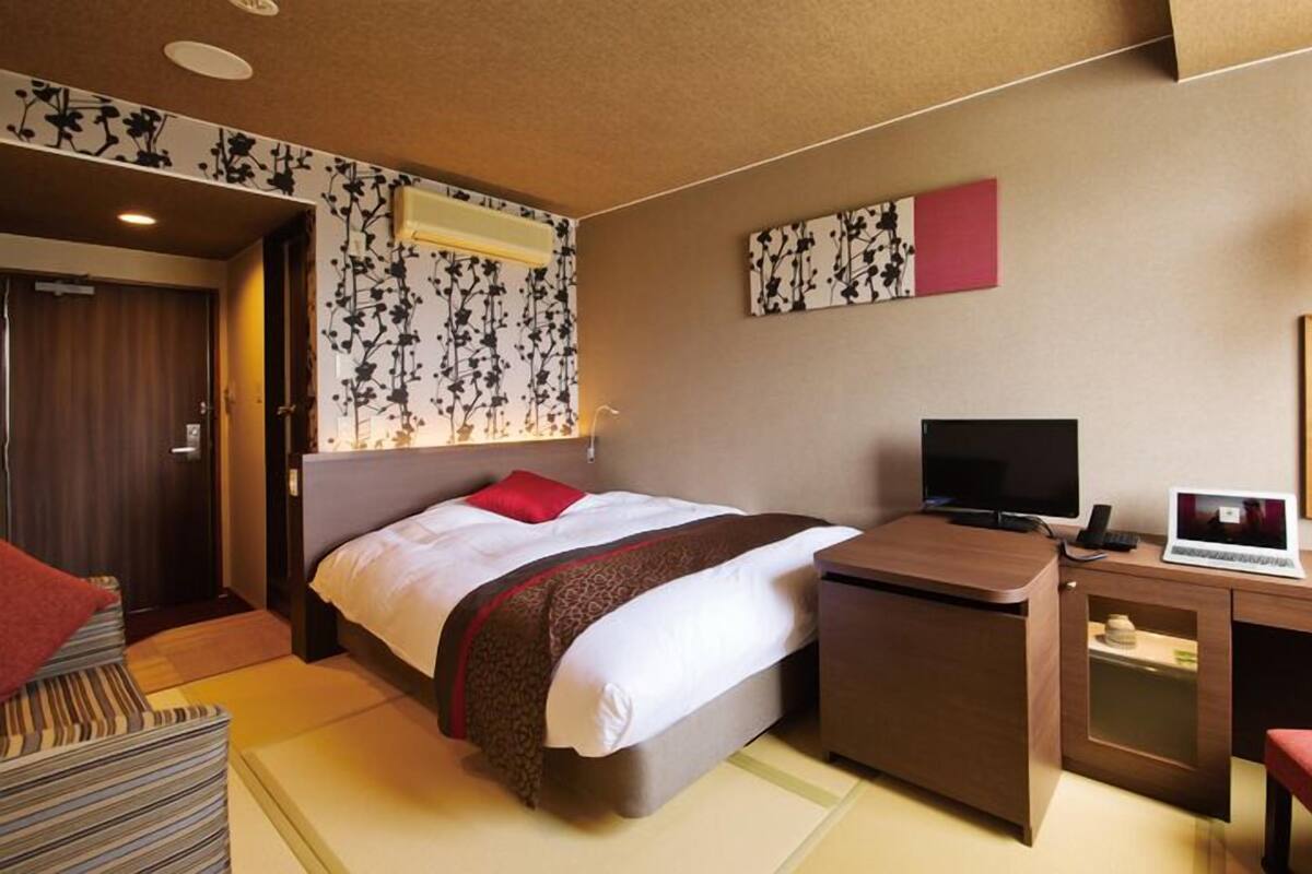 Habitación de hotel con cama, escritorio, silla, televisor y espejo.