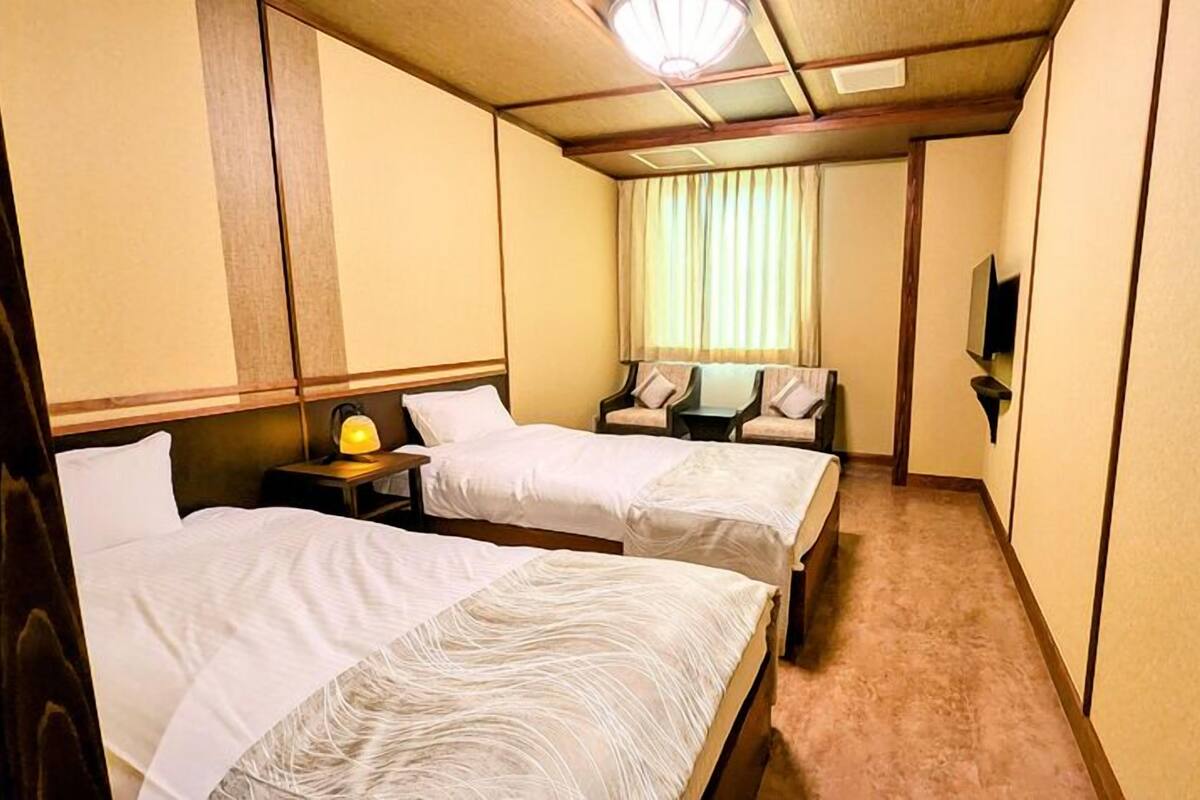 A hotel room with two beds, a nightstand with a lamp, a television, and a window with curtains.