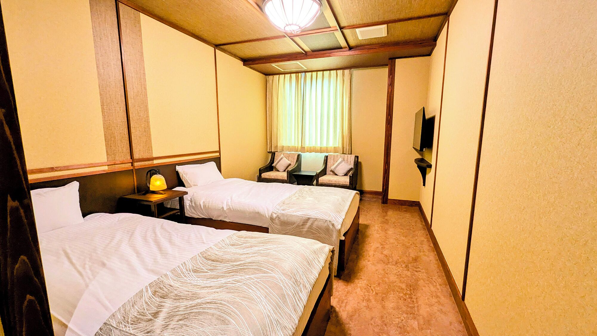 Una habitación de hotel con dos camas, una mesita de noche con lámpara, una pequeña mesa y un televisor en la pared.